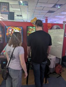 A person standing very close to their opponent's left side while they play pinball. 