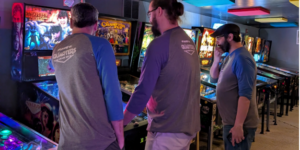 Two people crowd around one person playing pinball during a tournament.