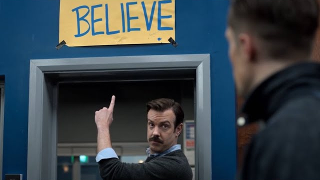 Ted Lasso pointing to a sign that says BELIEVE while giving a look to a player that says he means it. 